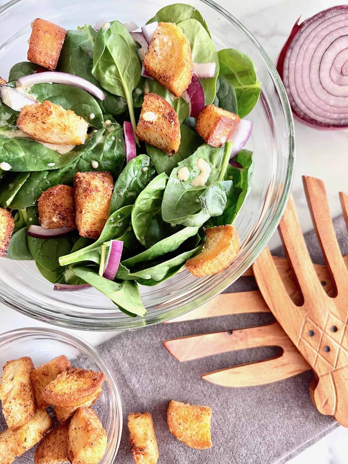 Simple Spinach Salad in a glass bowl next to ingredients of red onion croutons and cloth napkin and wood salad forks.