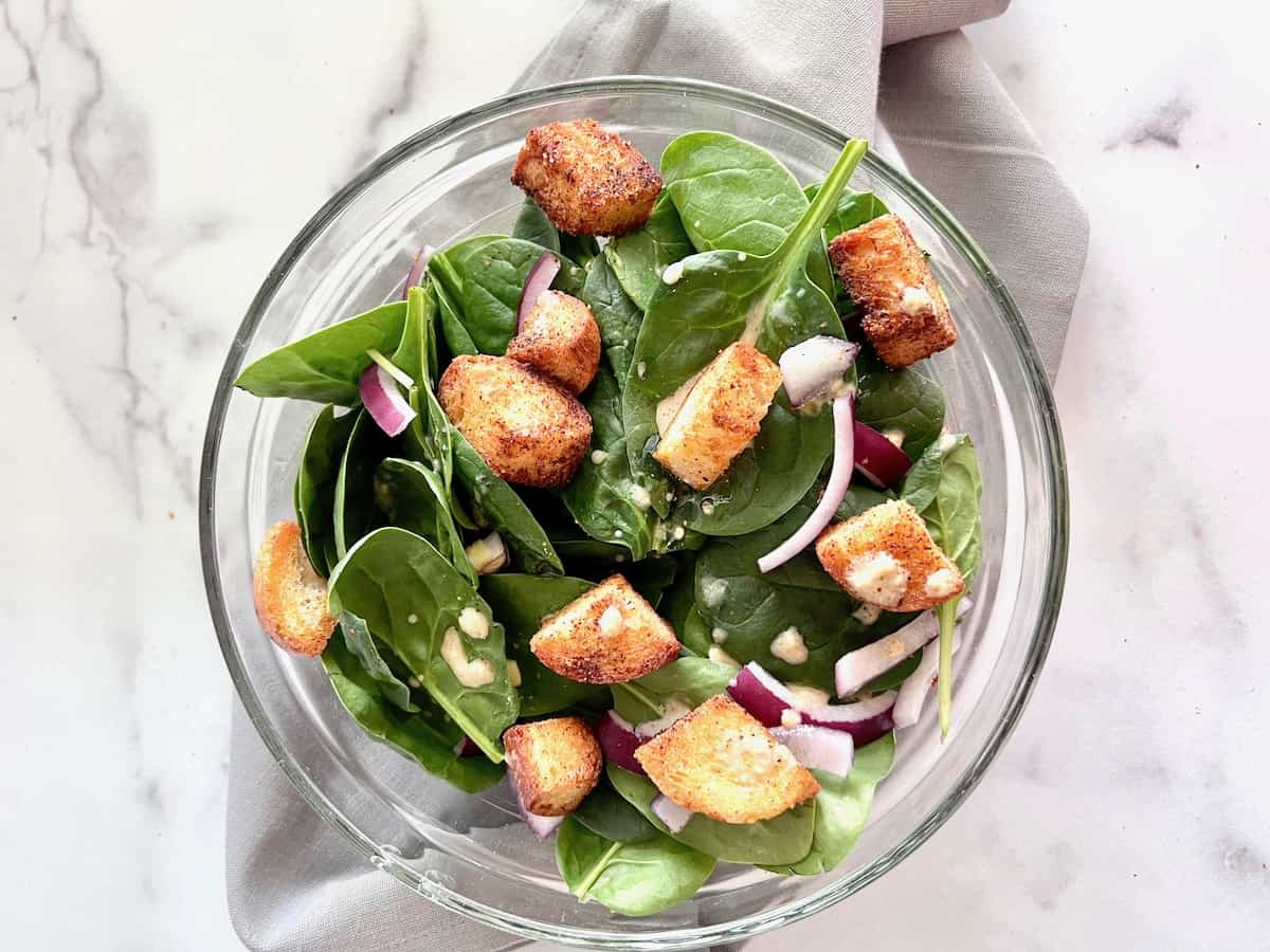 Landscape image of glass bowl filled with a spinach salad with onion, croutons, & a dijon vinaigrette.