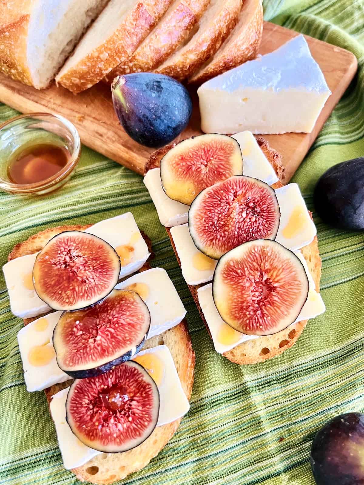 Fig and Brie Bruschetta On a green linen napkin next to cutting board with sliced bread figs cheese wedge and bowl honey.