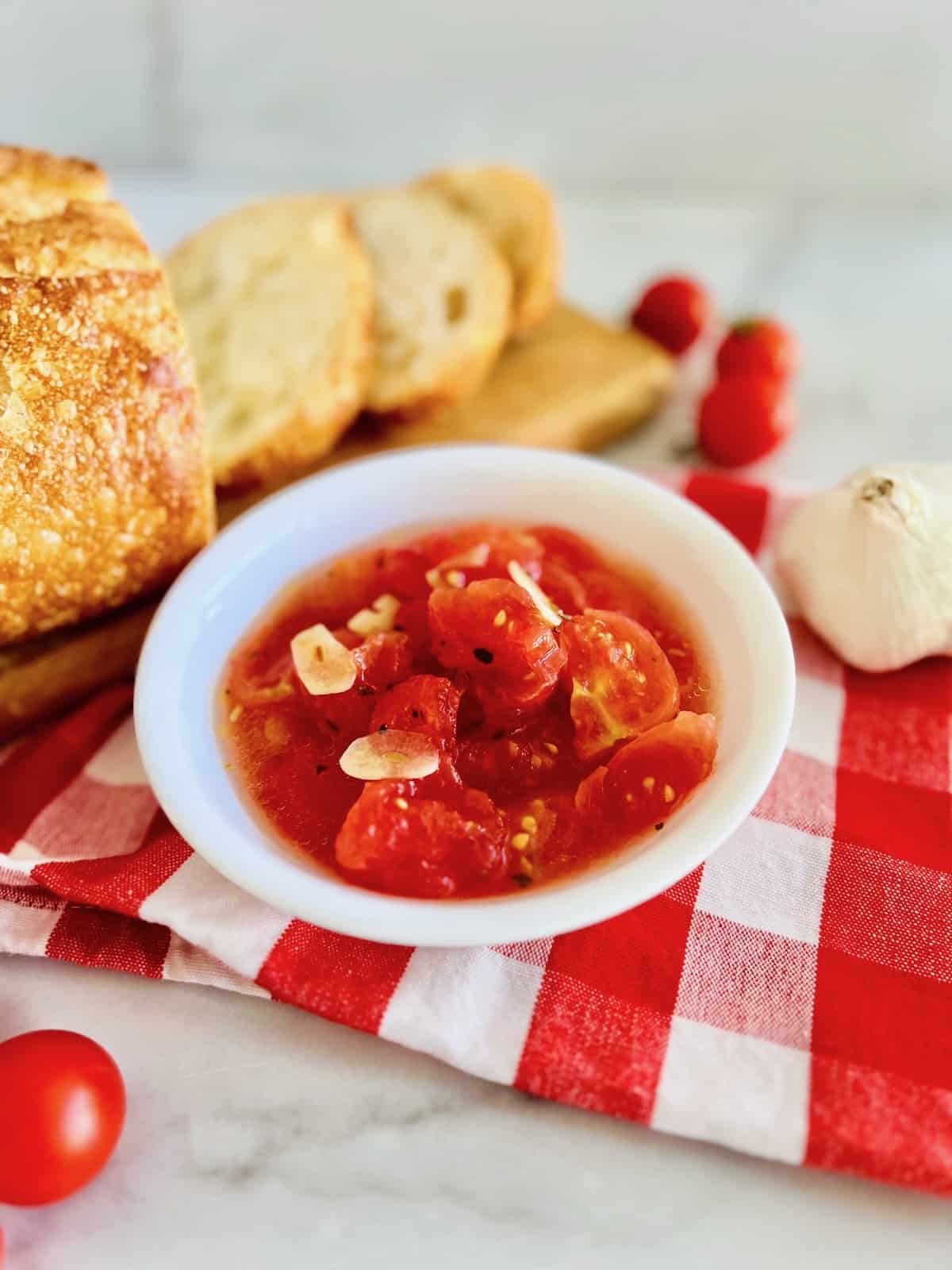Italian Marinated Tomatoes in a serving bowl next to a cutting board with freshly sliced bread for dipping or topping.