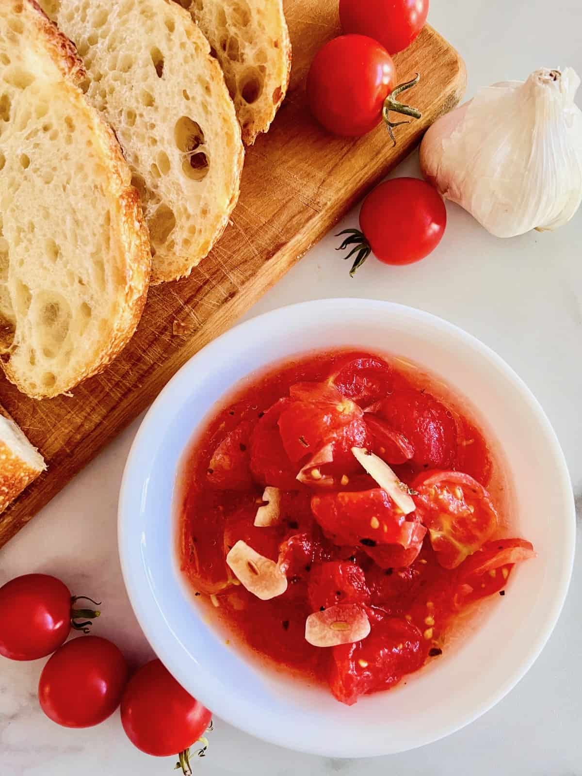 Italian Marinated Tomatoes in a bowl ready to serve with sliced bread on a board next to whole tomatoes and garlic bulb.