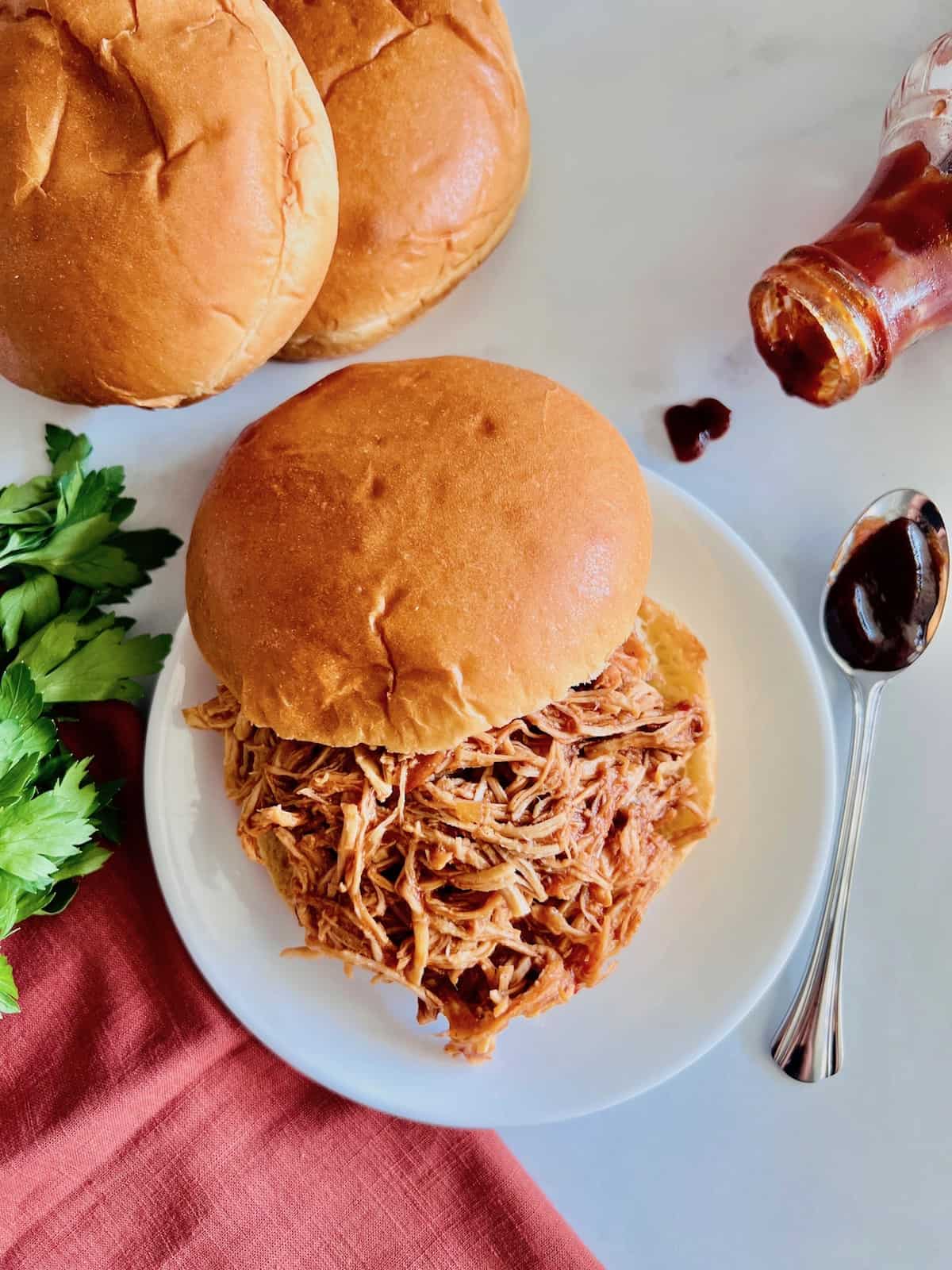 Slow Cooker BBQ Pulled Chicken On a bun plated next to buns bbq sauce in a bottle and on a spoon with parsley and red napkins.
