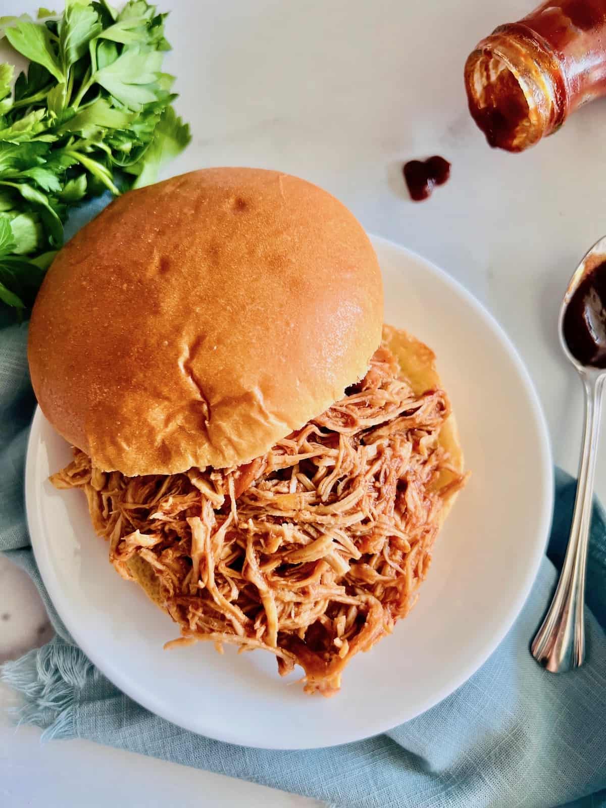 Slow Cooker BBQ Pulled Chicken On a bun with parsley and bbq sauce on the side with blue napkin.