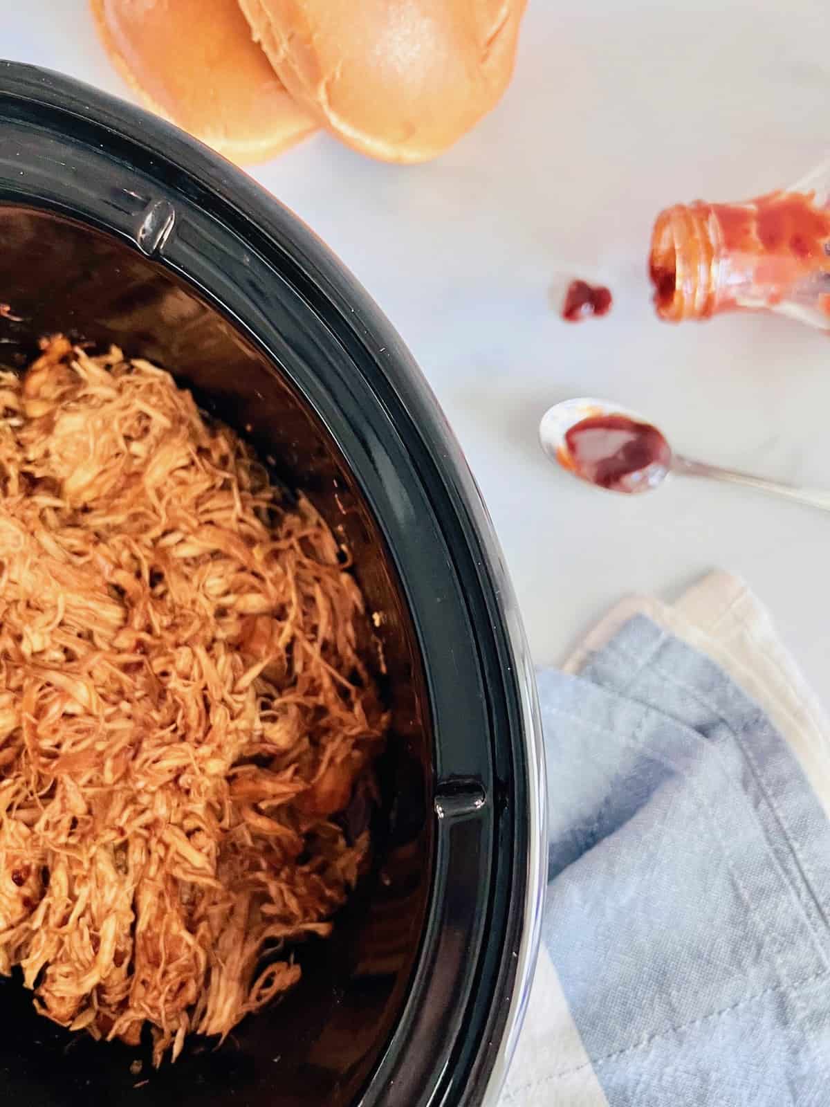 Slow Cooker BBQ Pulled Chicken Overhead in the crock next to bbq sauce and buns.