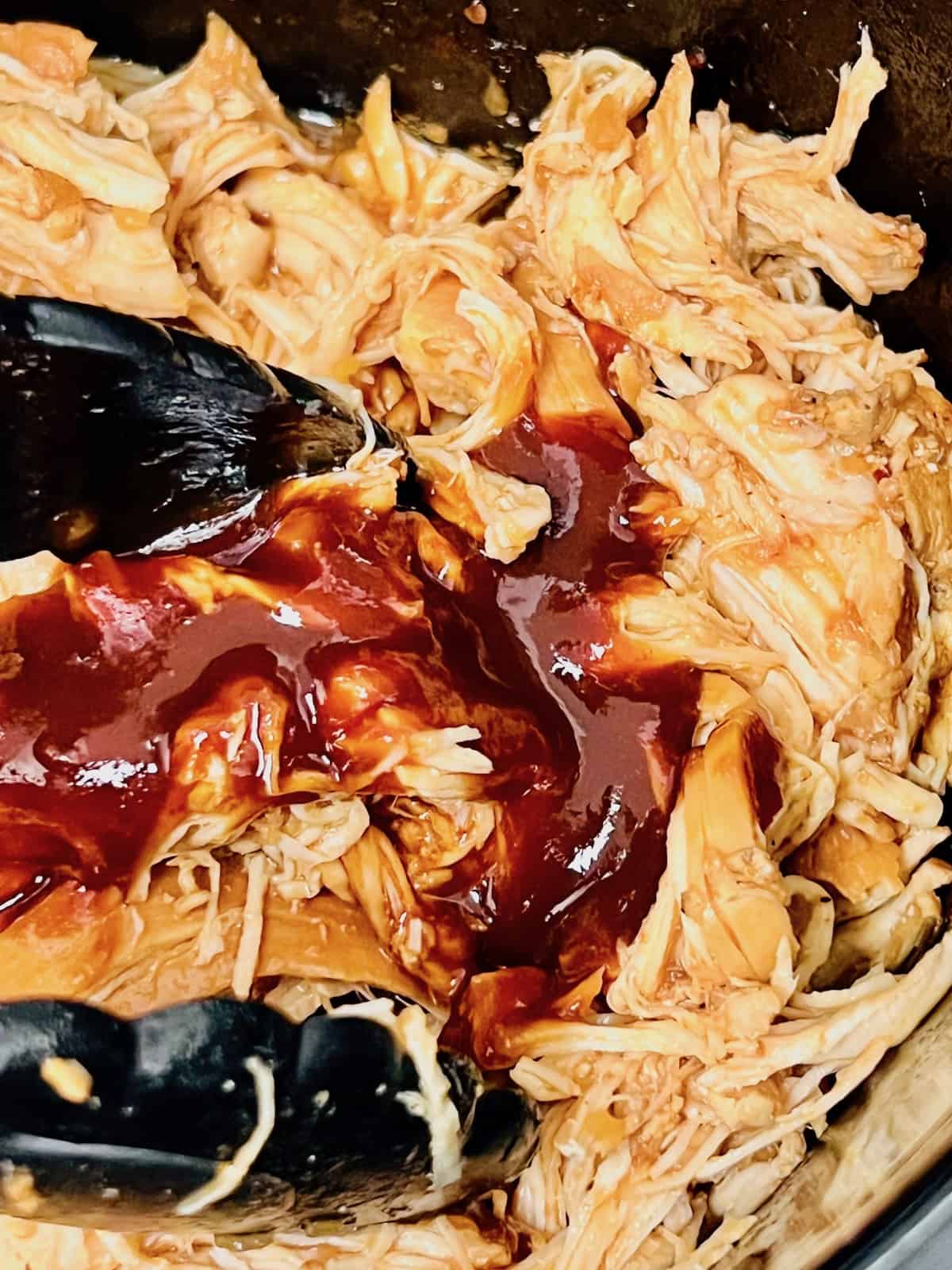Tongs mixing bbq sauce in with shredded chicken in Crockpot