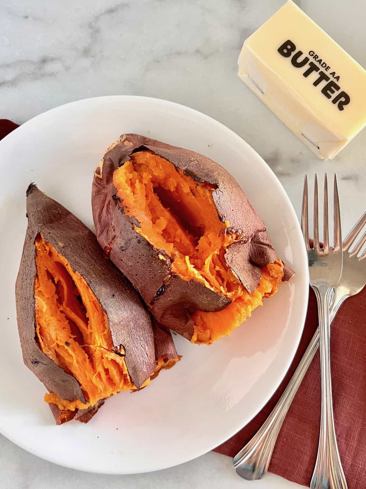 Air Fryer Baked Sweet Potatoes Split open on a plate next to forks and stick of butter.