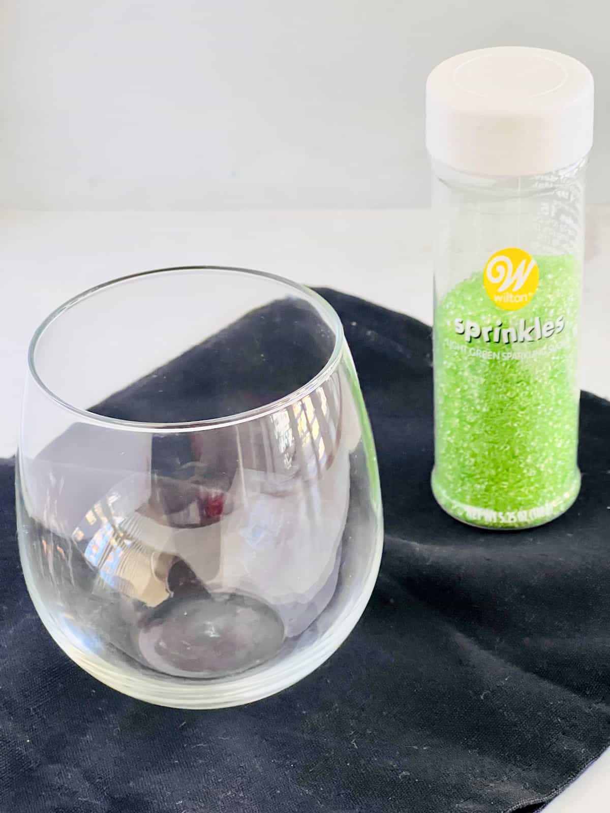 A round stemless wine glass next to a bottle of green sugar sprinkles.