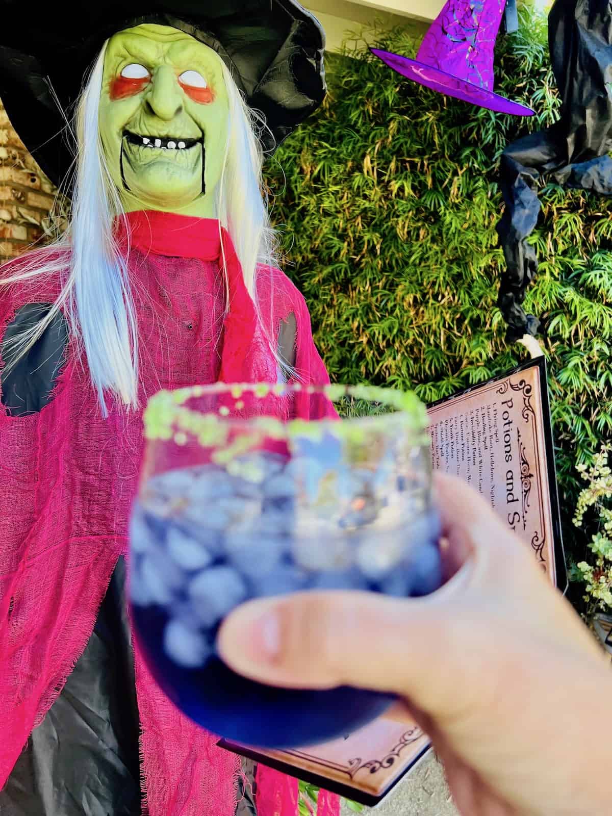 Witches Brew Cocktail in a glass being held in front of a decorative witch.
