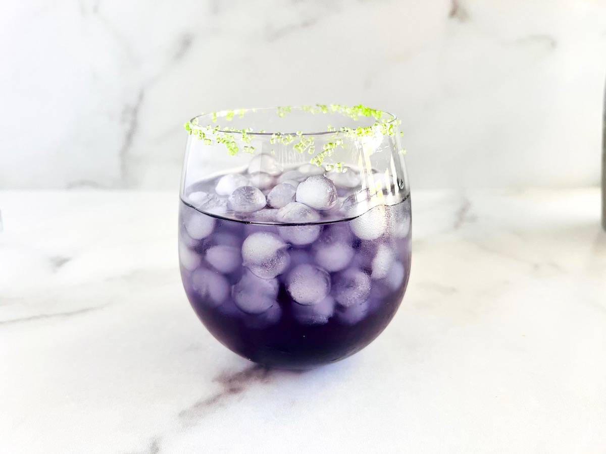 A glass of chilled purple drink is filled with ice cubes, sitting on a white marble surface. The rim of the glass is garnished with green sugar crystals, adding a contrasting pop of color.