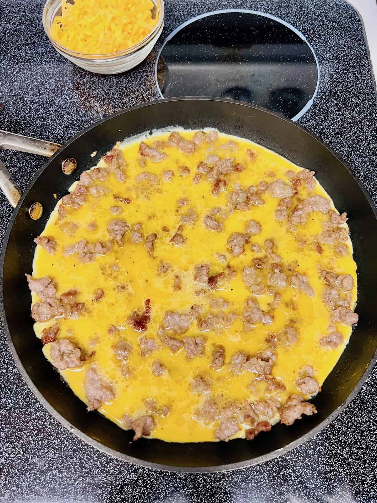 Liquid eggs in the skillet with cooked sausage crumbles.