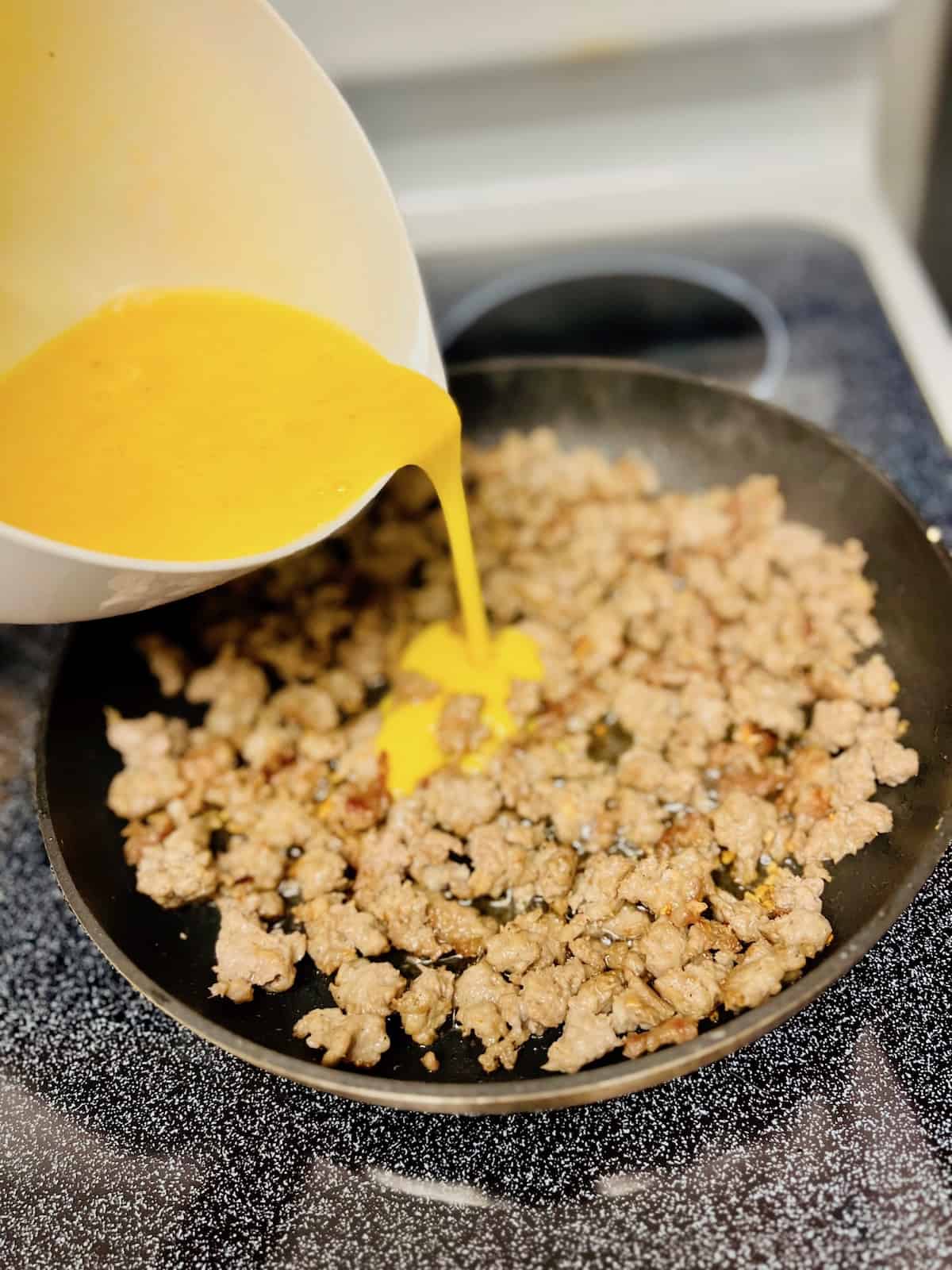 Pouring raw scrambled eggs from bowl into pan with breakfast sausage.