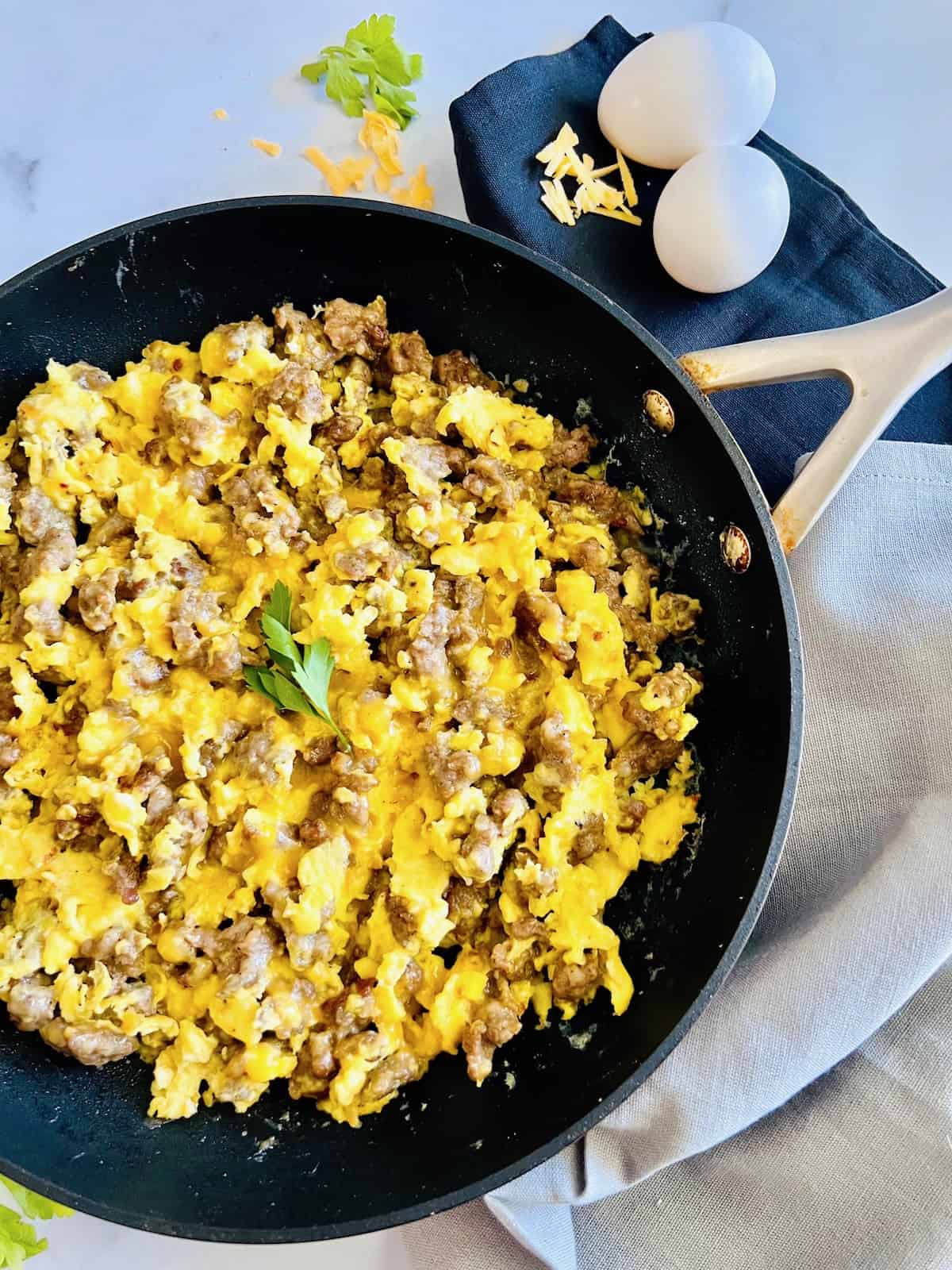 Breakfast Sausage, Egg, & Cheese Scramble in a skillet topped with parsley leaves next to eggs in the shell and shredded cheese.