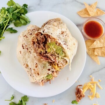 Burrito filled with pork carnitas and loaded with cheddar cheese, mexican rice, refried beans, sour cream, fresh avocado, cut in half on a plate next to cilantro, hot sauce, and chips.