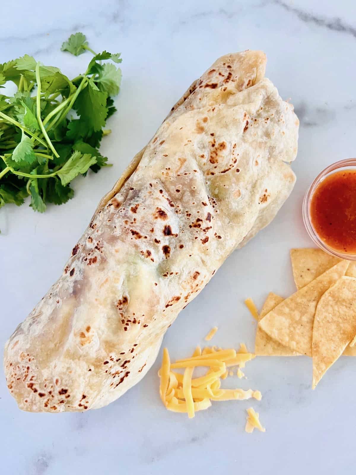 Carnitas Burrito Wrapped up on the counter next to hot sauce chips cheese and cilantro.