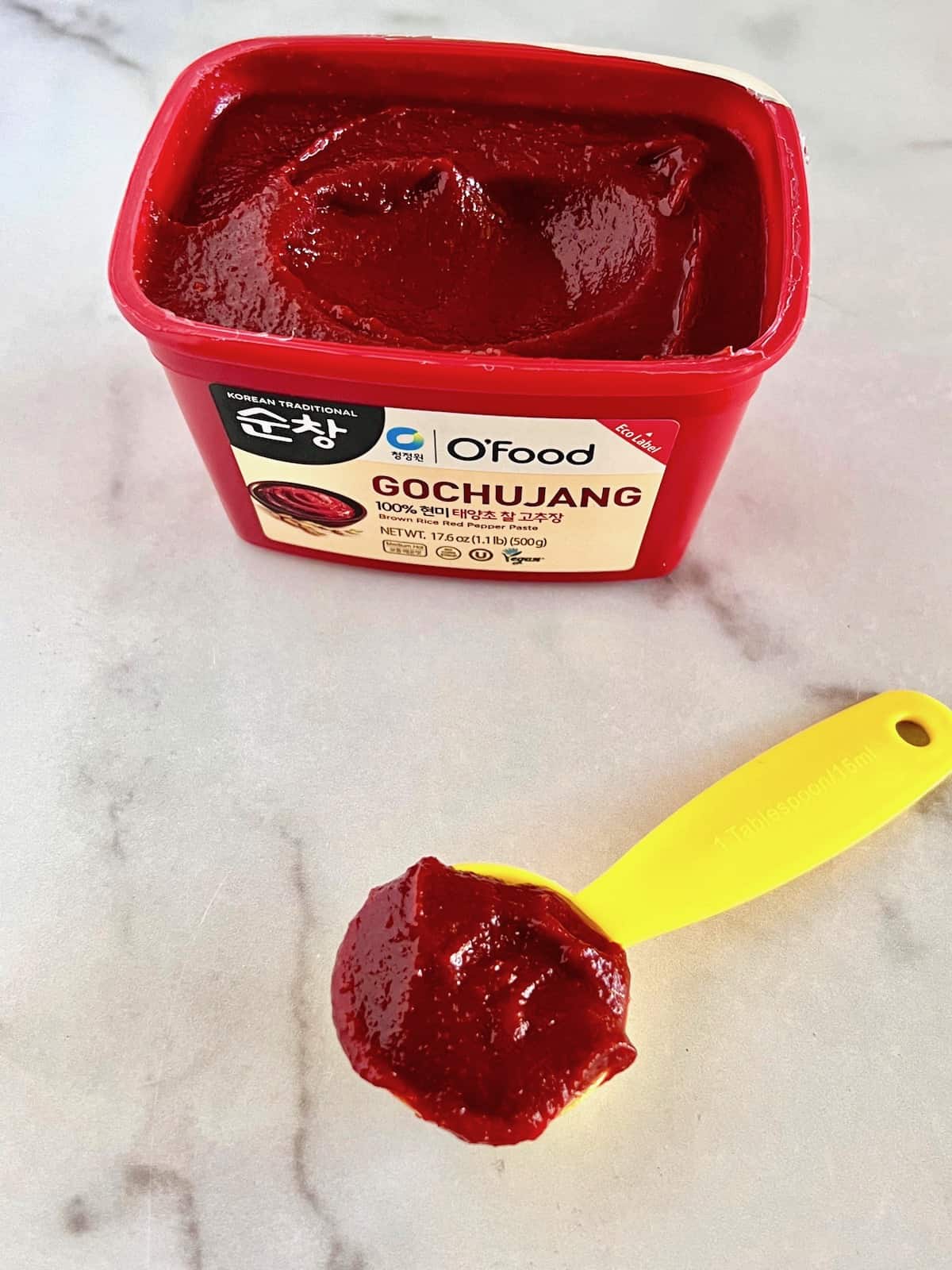 Tub of gochujang next to a measuring tablespoon filled with the spicy korean paste.