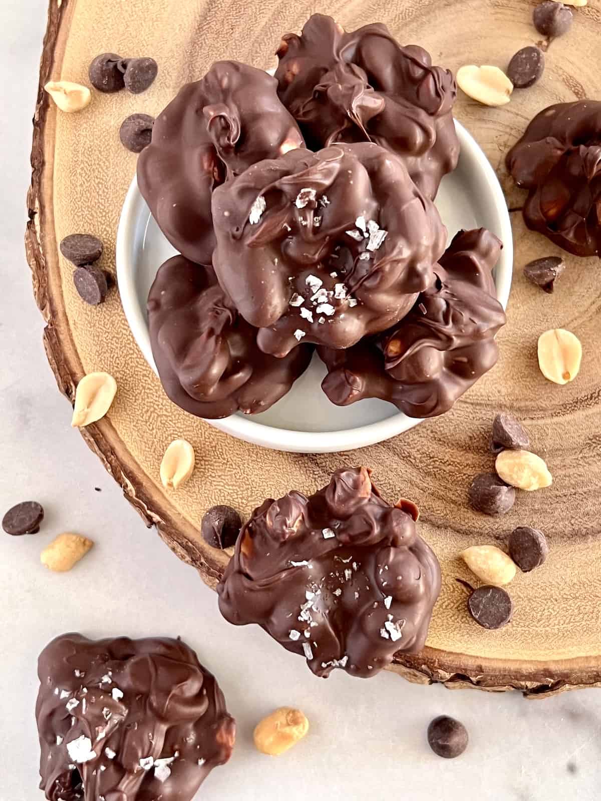 Chocolate Peanut Clusters In a bowl on a wood plank with clusters peanuts and chocolate chips surrounding it.