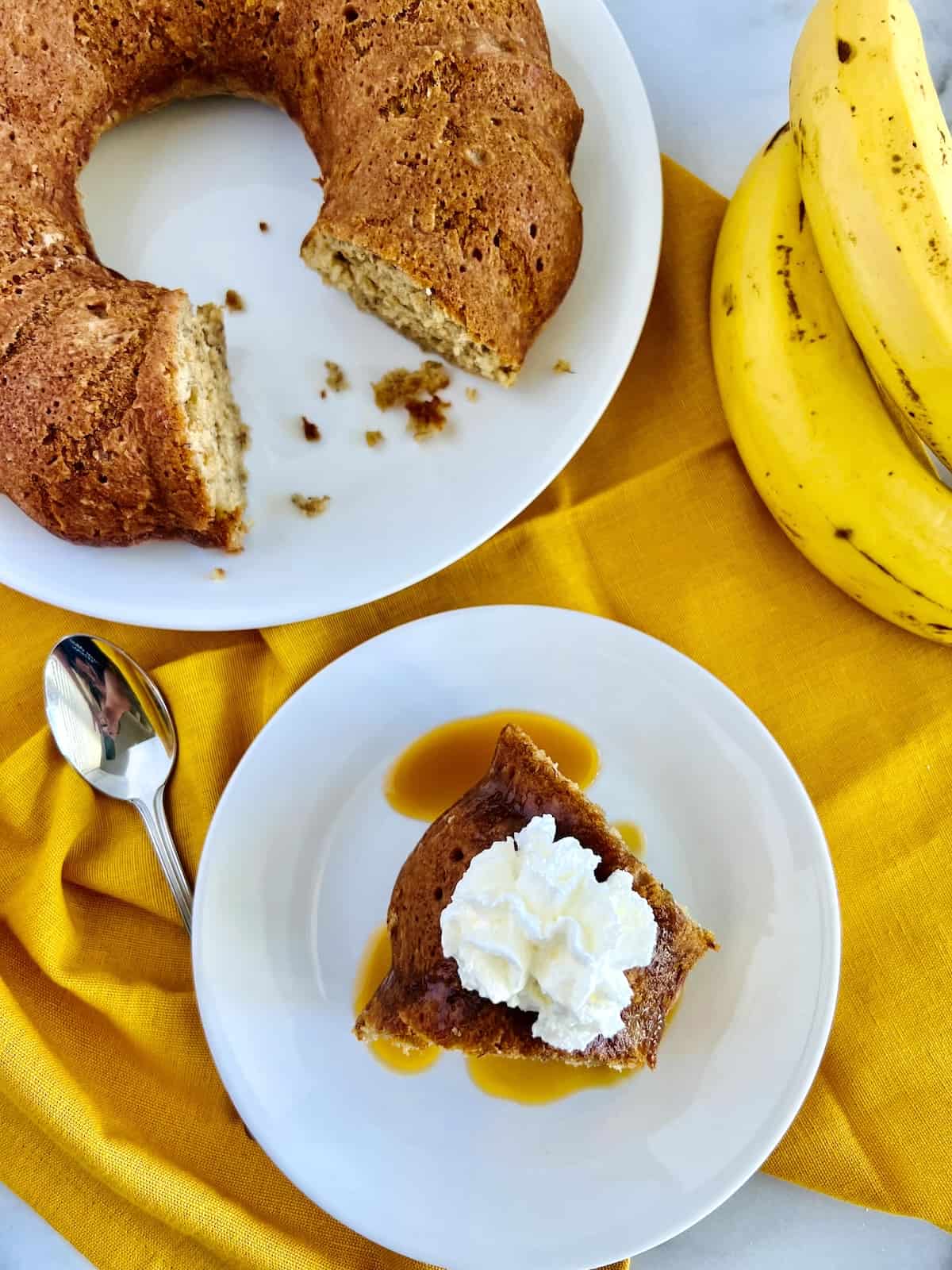 Banana Bread Bundt Cake Recipe done and plated with caramel sauce whipped cream next to spoon cake plate and bananas.