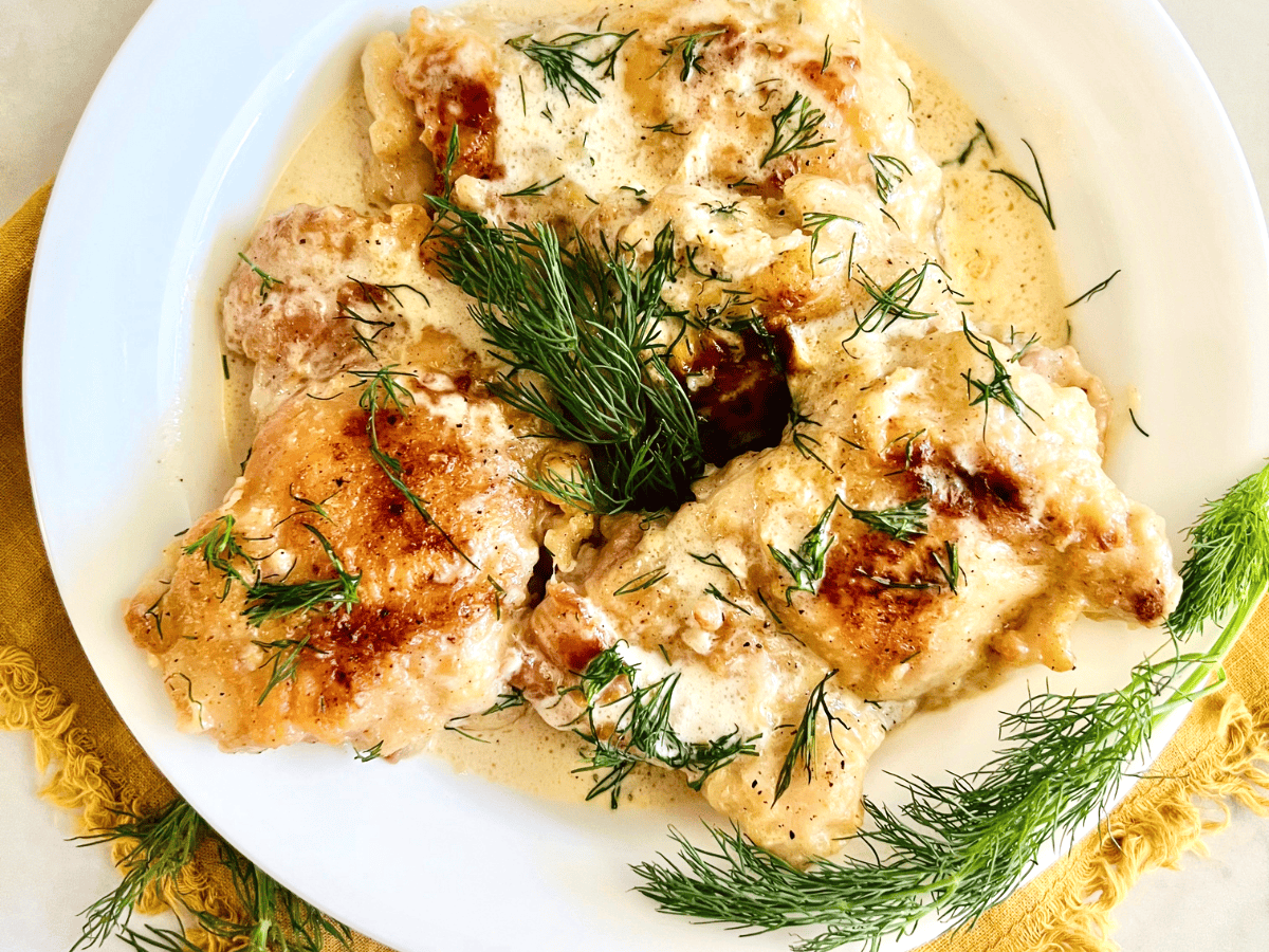A white plate features crispy chicken thighs in a creamy dill sauce garnished with fresh dill sprigs. The dish sits on a yellow fringed napkin, adding a pop of color to the presentation. The chicken is golden-brown and looks tender and flavorful.