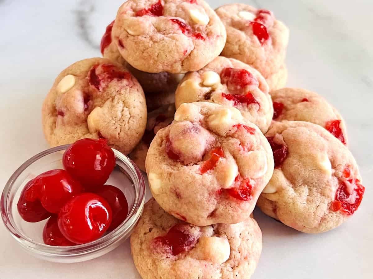 Cherry White Chocolate Chip Cookies stacked next to bowl of maraschino cherries.