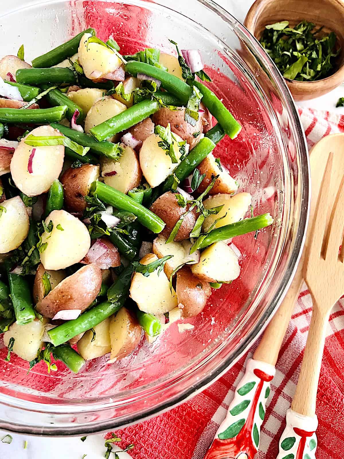 Italian Green Bean and Potato Salad Serving bowl with wooden utensils and bowl of fresh herbs.