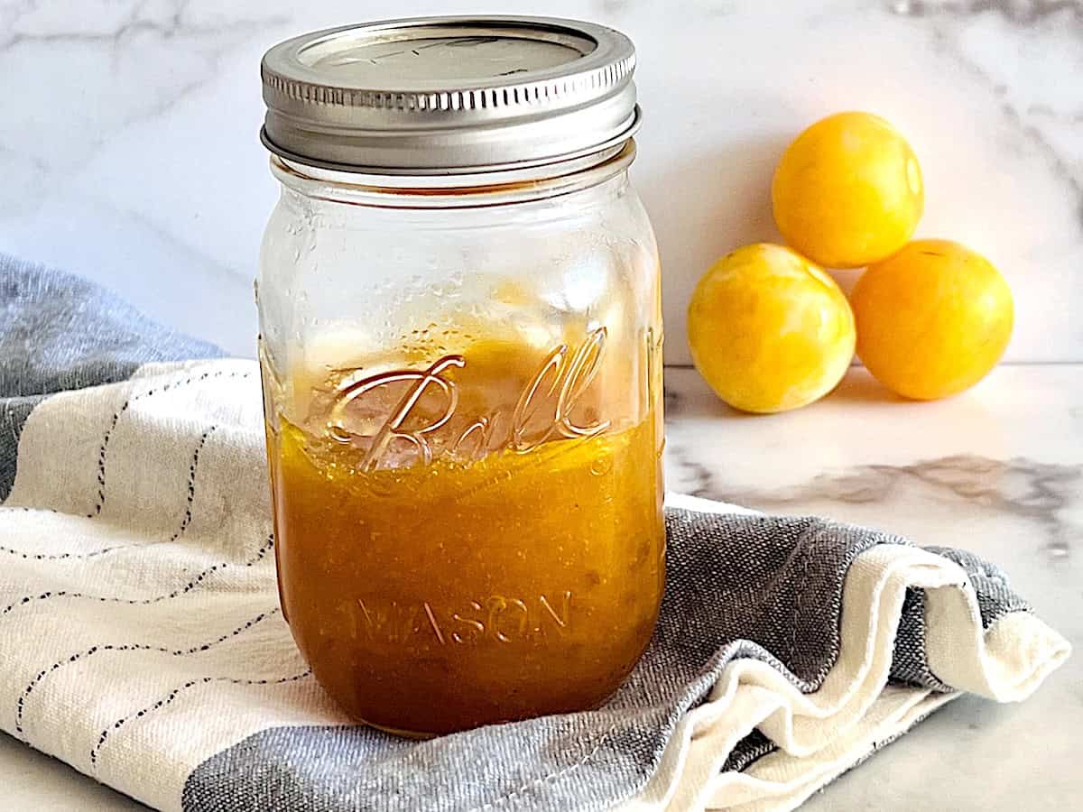 Jar of plum jam next to fresh fruits on a cloth napkin.