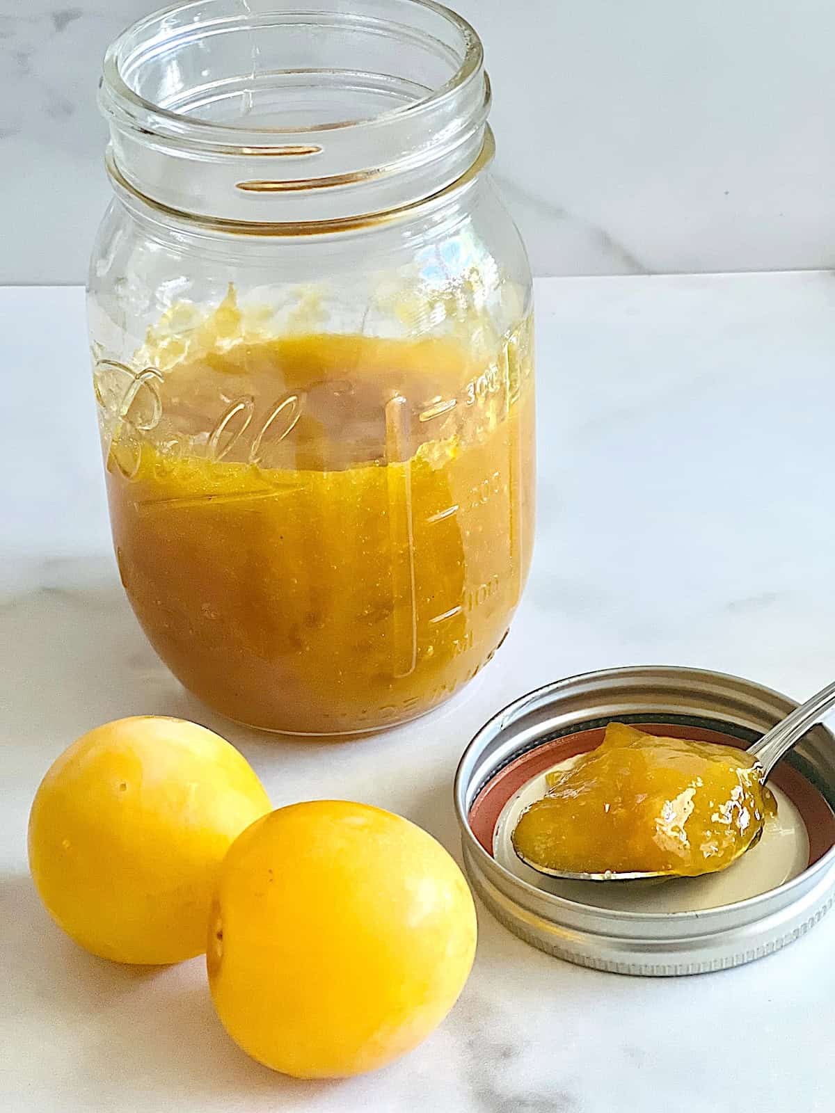 Plum Jam open jar with spoonful on the lid next to whole fresh yellow plums.