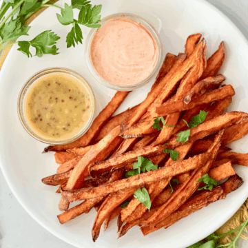 Crispy sweet potato fries with dipping sauces on a white plate, garnished with parsley. Perfect for a tasty snack or side dish.