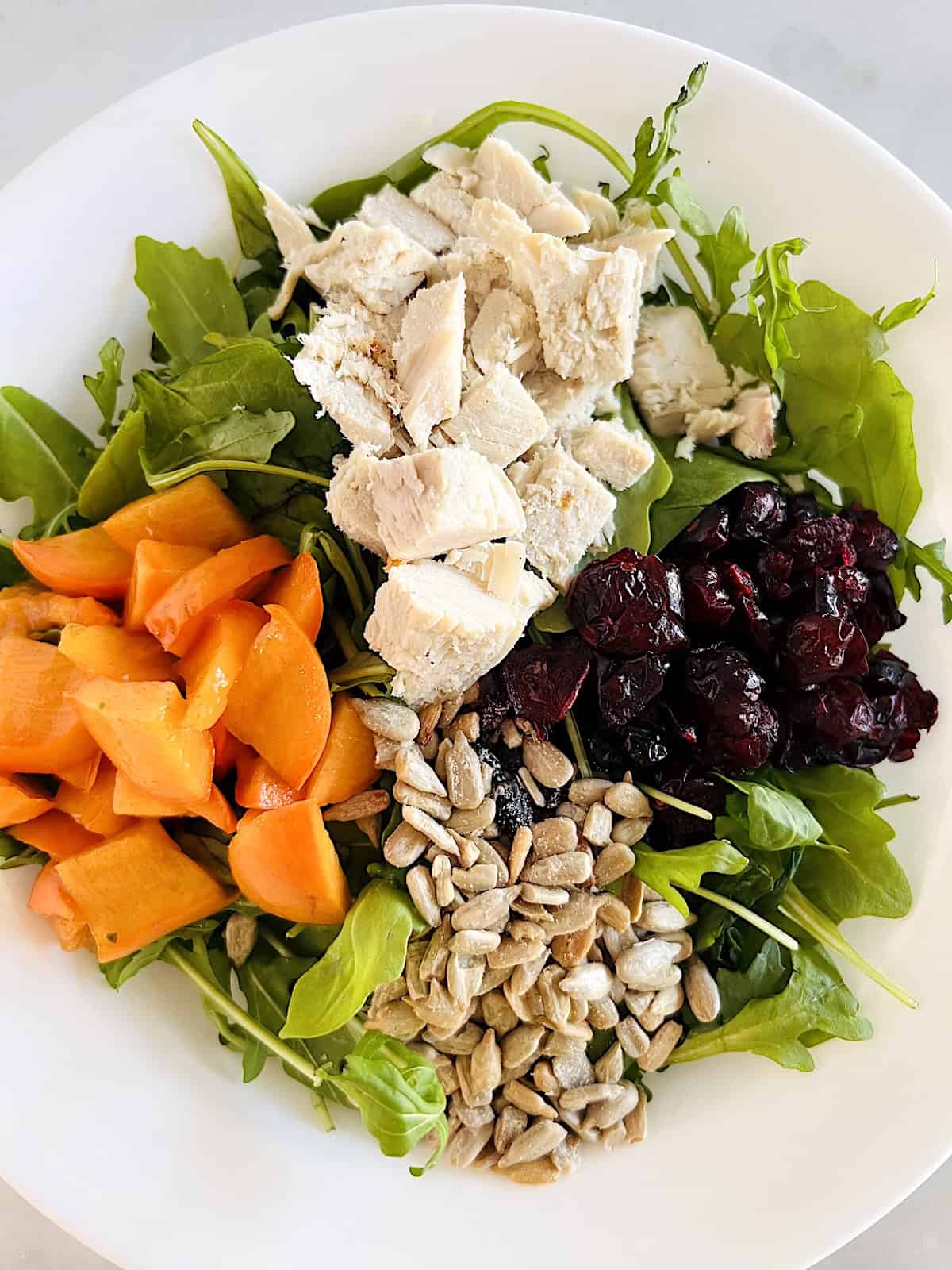 Winter Green Salad with Turkey Arugula and spinach topped with diced turkey persimmon dried cranberries and sunflower seeds in a bowl.