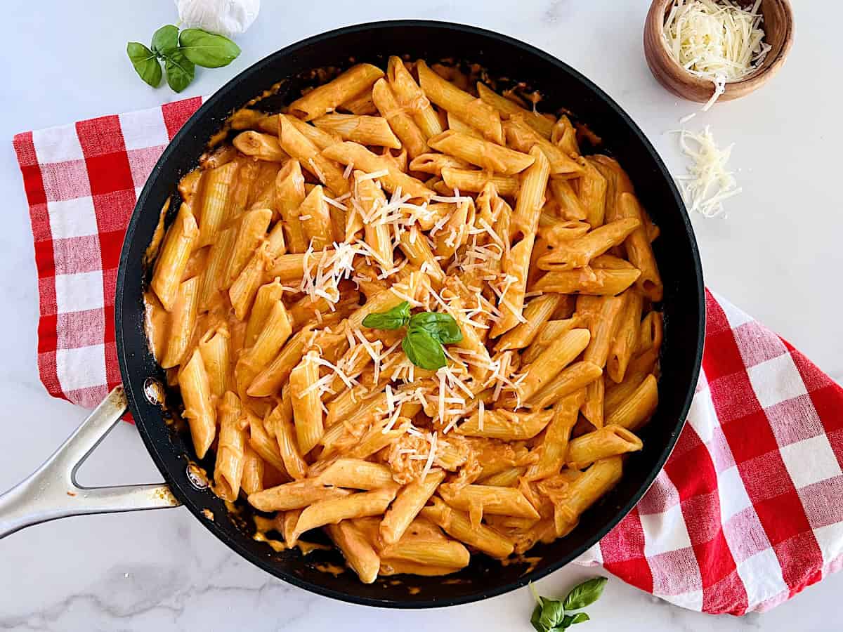 Red checkerboard napkin on the table with a skillet of homemade penne pasta alla vodka topped with parmesan cheese and fresh Italian basil leaves.