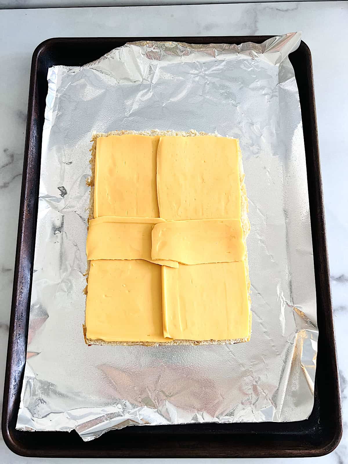Baking sheet with bottom of rolls topped with layer of American cheese slices.