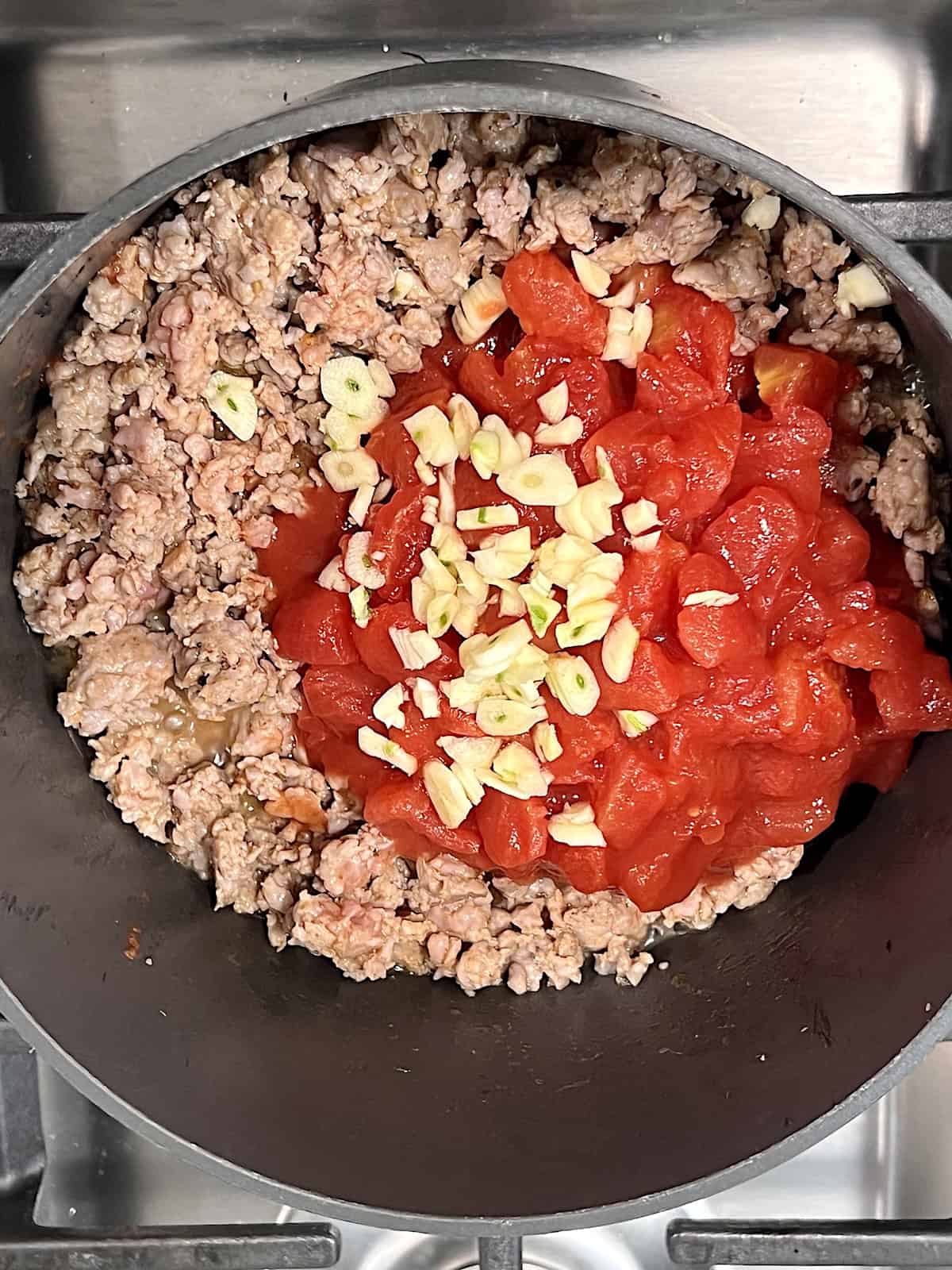 Adding diced tomatoes sliced garlic to pot with cooked ground meat.