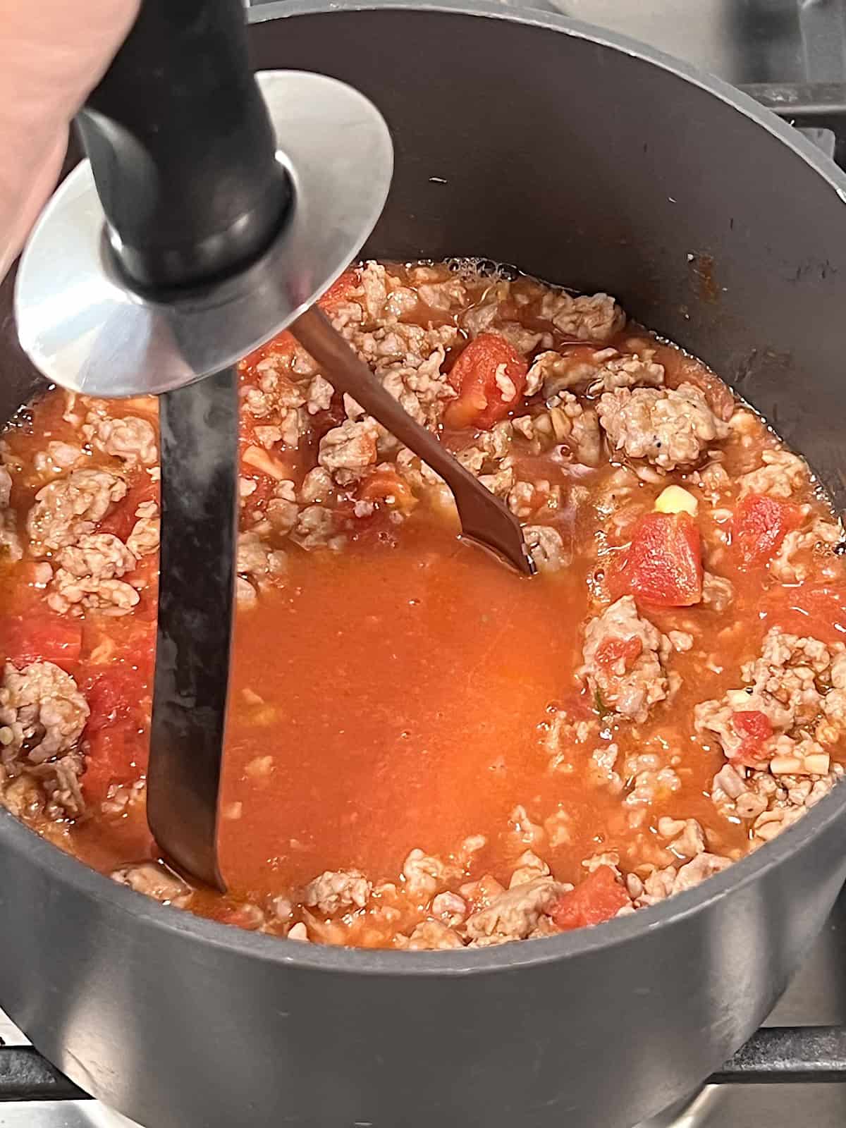 using a potato masher to break up sausage in the tomato sauce/