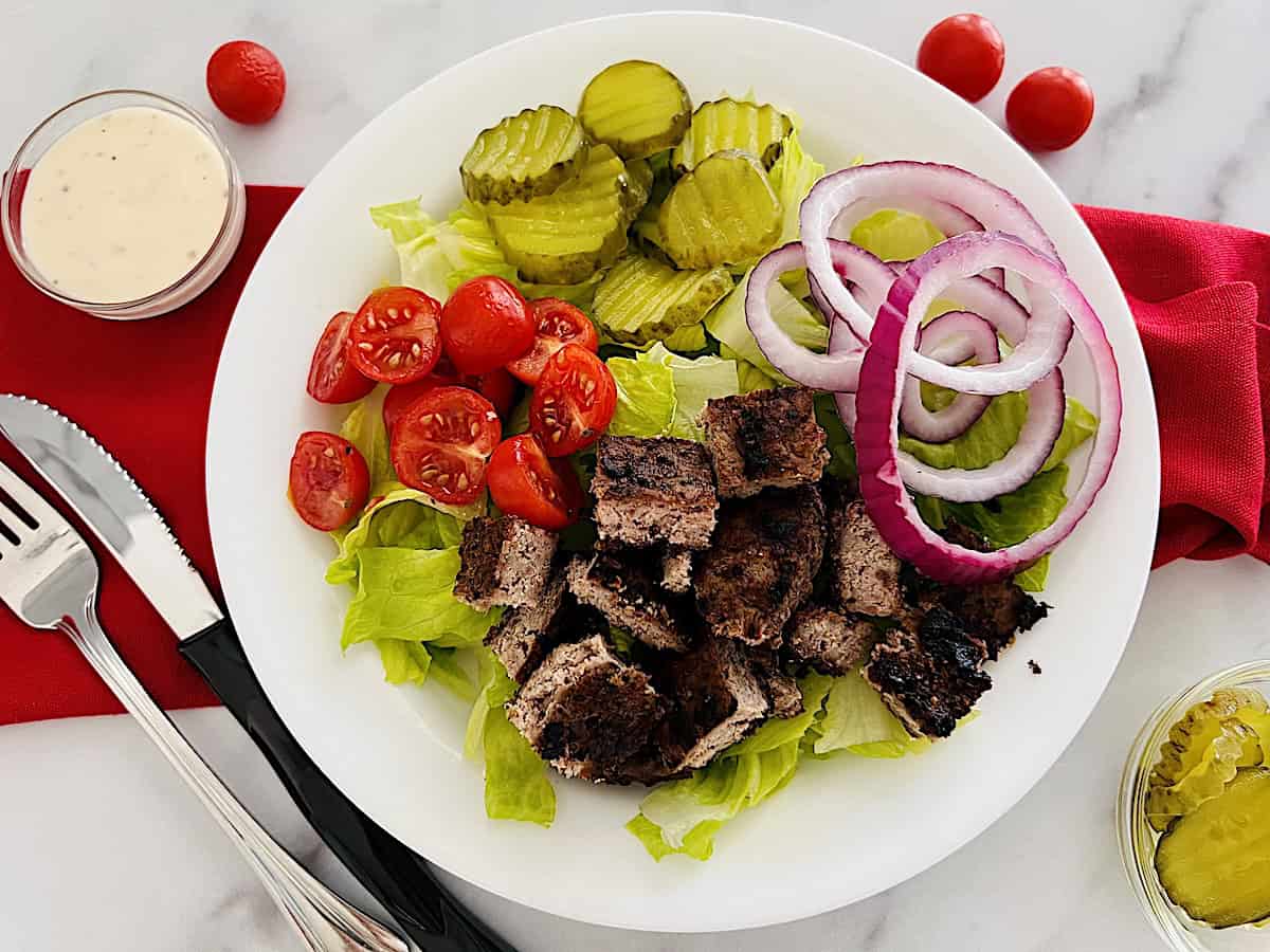 Healthy hamburger protein Bowl meal with ground beef, romaine, tomatoes, red onion slices and dill pickles.