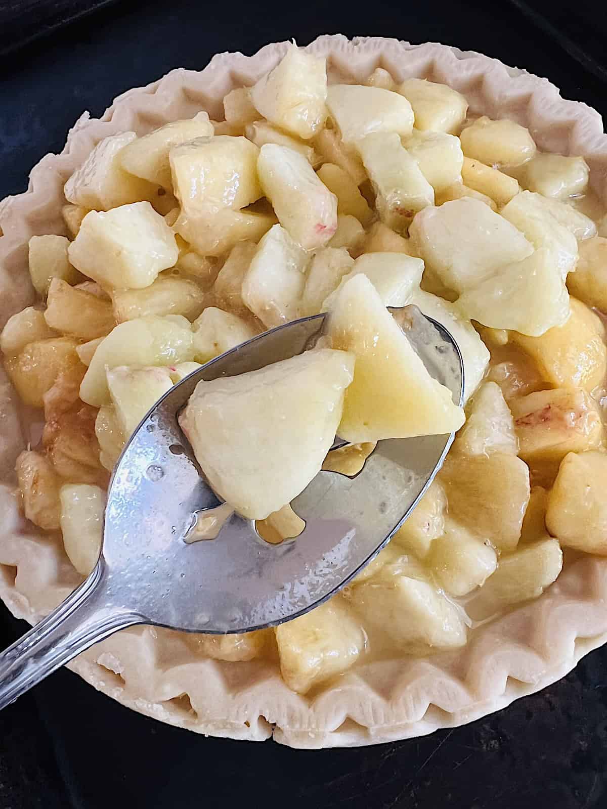 Slotted spoon adding fresh peach chunks to pie crust.