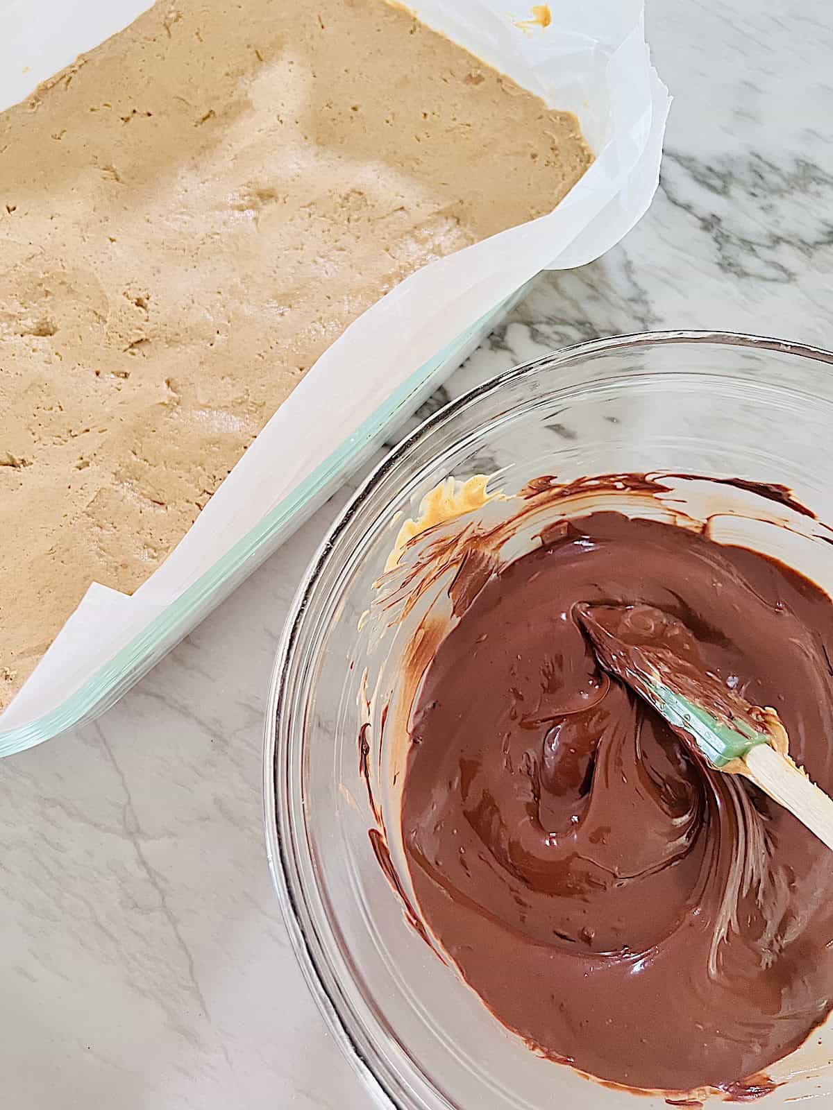 No Bake Chocolate Peanut Butter Bars Pan with pb layer next to bowl of melted chocolate.
