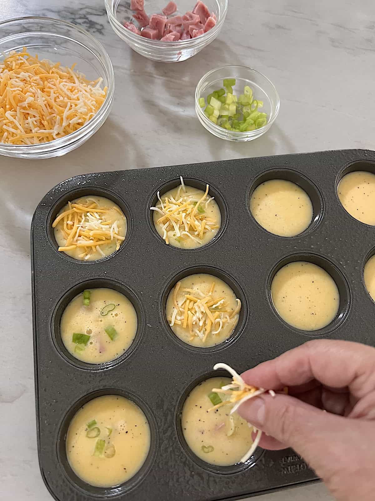 Mini Egg Bites Adding cheese ham and green onion to egg mixture in muffin tin.