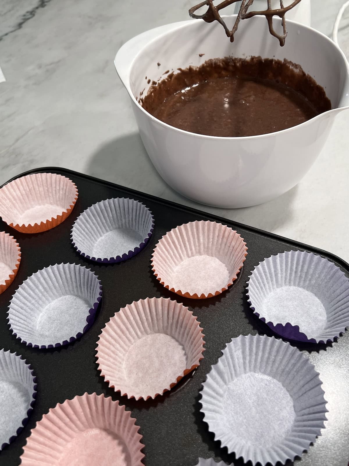 Bowl with chocolate cake batter next to cupcake liners in a pan.